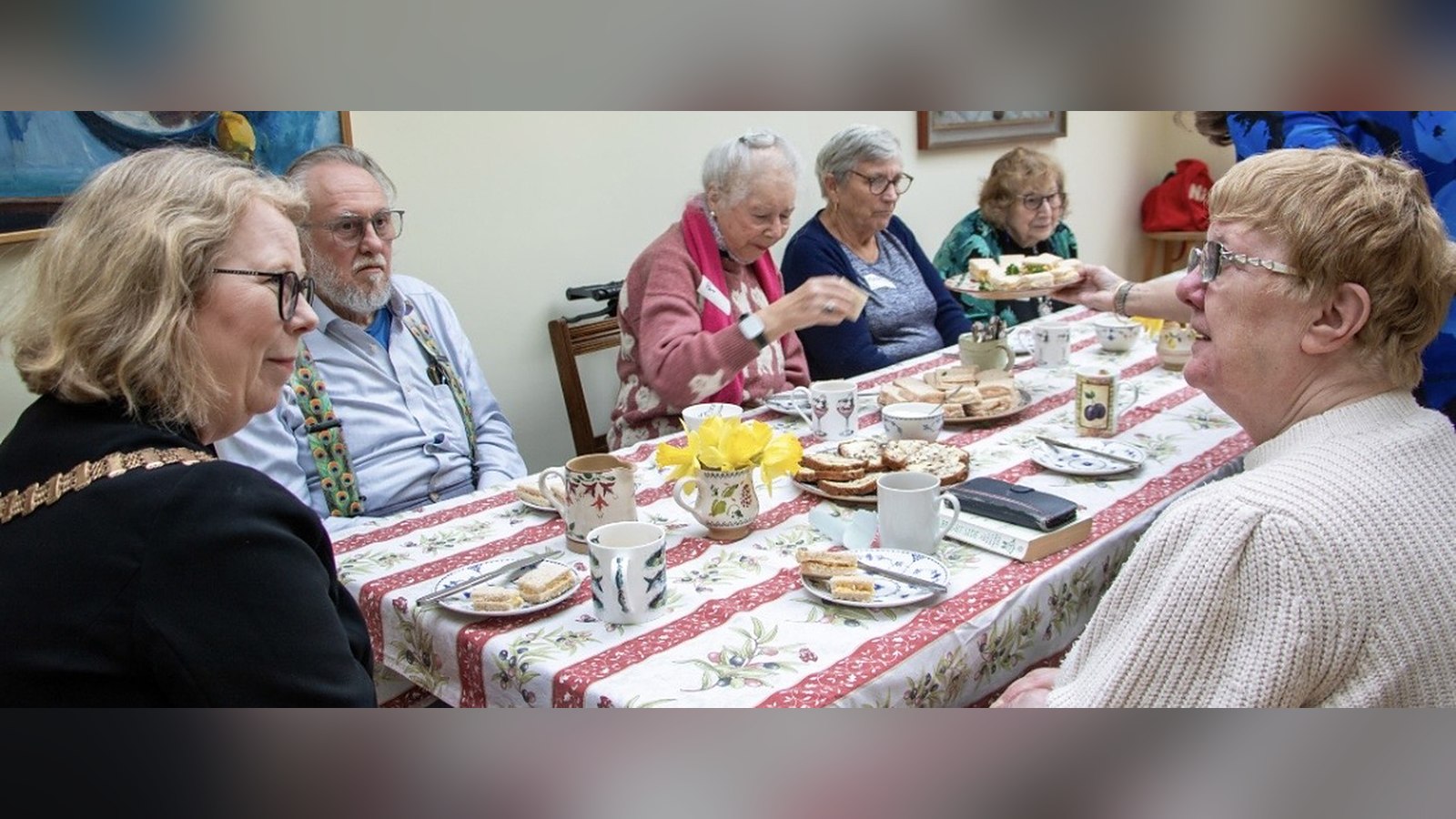 Personas mayores conversando alrededor de una mesa con té y pasteles